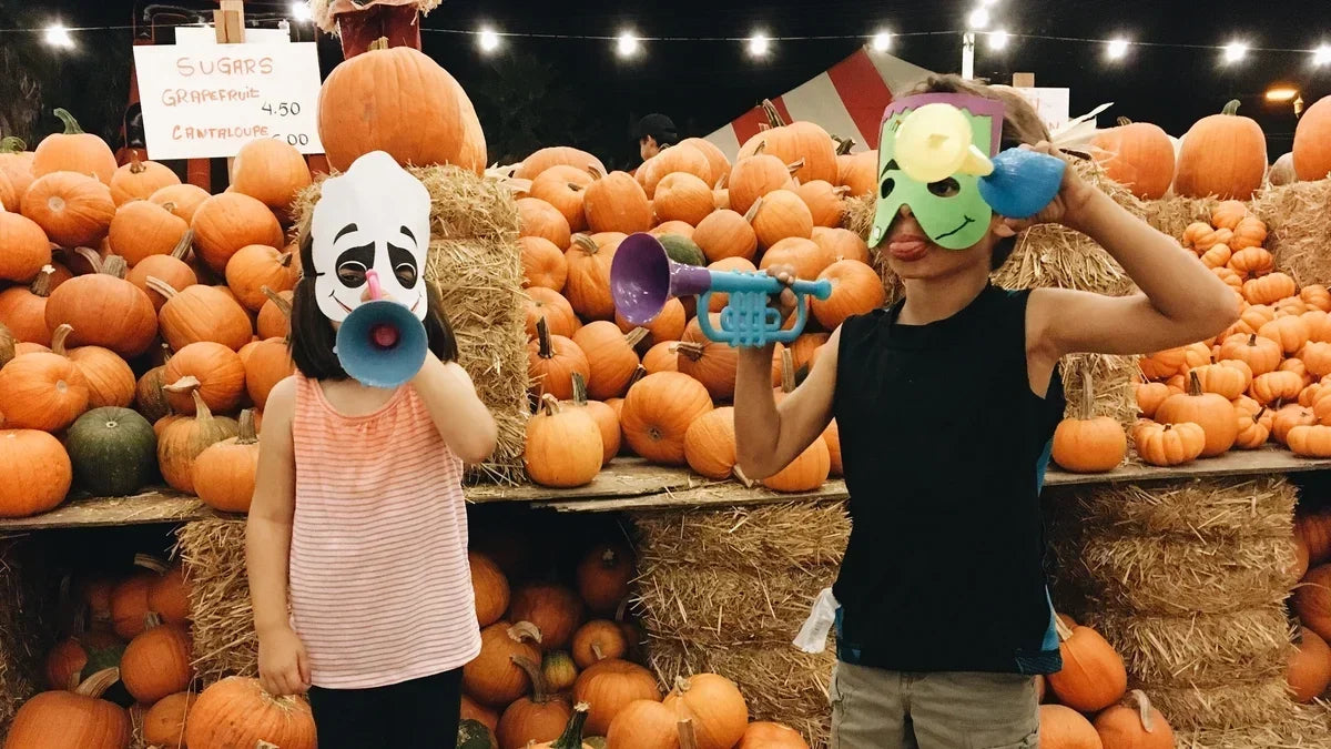 Two children standing in front of a lot of pumpkins wearing halloween masks. 